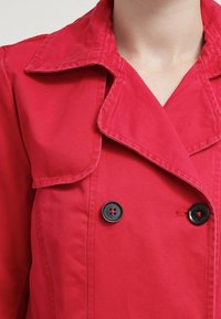 Abrigo rojo de tipo trench con un cuello ancho, diseño de doble botonadura y botones negros. Textura de tela lisa con detalles de costuras visibles.