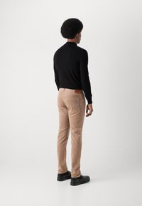 Cămașă neagră cu mâneci lungi, pantaloni din cord dintr-o nuanță deschisă de maro, vedere din spate. Pantalonii au buzunare pe spate și un material texturat; încălțăminte netedă.