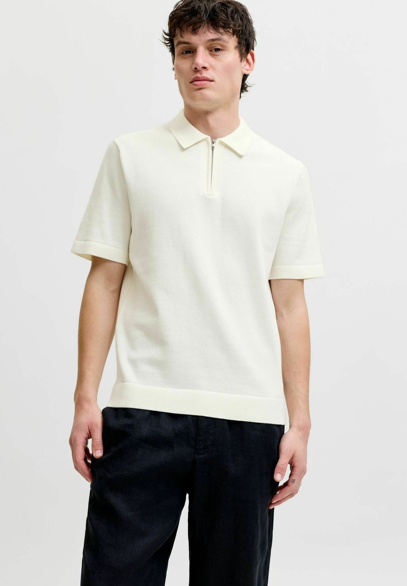 Jeune homme portant une chemise blanche à manches courtes avec col zippé et un pantalon noir ample, debout devant un fond gris uni.