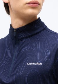 Κοντινό πλάνο ατόμου που φορά μπλε ναυτικό τοπ με φερμουάρ, διακριτικό στροβιλιστό μοτίβο και λευκό λογότυπο Calvin Klein στο στήθος.