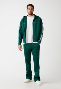 Tuta verde con cappuccio zip e pantaloni a fantasia, con strisce bianche lungo le maniche e i lati, abbinato a sneakers bianche.