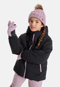 Fille portant une veste d'hiver noire, un bonnet en tricot lavande avec un pompon, des gants assortis et un pantalon de neige lavande sur fond blanc.