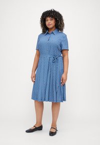 Mėlyna trumpomis rankovėmis suknelė su gėlėmis, mygtukų priekyje, juosmenį akcentuojanti ir susiraukšlėjusi sijonu. Nešiojama su juodais plokščiais bateliais.