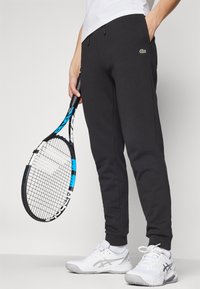 Sorte treningbukser med elastisk liv, sidelommer og en liten grønn logo. Kombinert med hvite tennissko og holder en racket med blå detaljer.