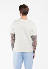 Lichtgrijze t-shirt met korte mouwen, gemaakt van zacht materiaal, met een relaxte pasvorm en een ronde hals. Opvallende tatoeages zichtbaar op de armen.