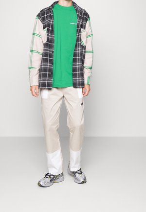 Zielona koszulka pod czarno-białą koszulą w kratę z zielonymi akcentami, w połączeniu z beżowymi spodniami z białymi panelami i szarymi trampkami.