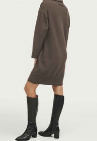 Person som bär en lång brun tröjdress och svarta knähöga läderstövlar, står framför en enhetlig ljus bakgrund.