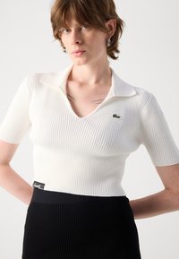 Weißes, geripptes Strickpolohemd mit V-Ausschnitt, kurzen Ärmeln und einem kleinen grünen Logo auf der Brust. Kombiniert mit einem schwarzen, gerippten Rock.
