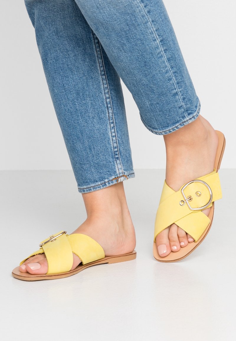 Pieds portant un jean bleu et des sandales plates jaunes avec des brides larges croisées et une grande boucle ronde sur un sol clair.