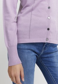 Cardigan tricotat de culoare violet deschis, cu un design ajustat, mâneci lungi și nasturi negri. Purtați-l peste blugi din denim albastru, evidențiind textura netedă.