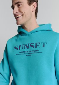 Turquoise hoodie met een buidelzak, voorzien van een grafische print in donkerblauw. De tekst luidt "ZONSONDERGANGSHORIZON IN DE AVOND" en "NO-EXCESS."