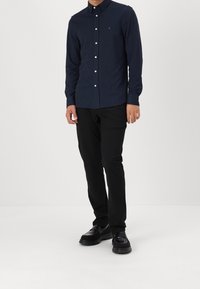 Námořnická košile na knoflíky s límečkem, vyrobená z texturované tkaniny, spárovaná s černými kalhotami a černými botami. Na košili je decentní logo.