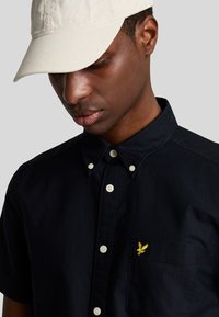 Camicia button-up blu scuro con taschino e logo ricamato giallo; abbinata a un cappellino beige. Tessuto morbido e colletto strutturato.