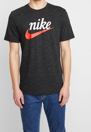 Mann trägt dunkelgraues Nike-T-Shirt mit rotem Swoosh-Logo und blauen Jeans vor weißem Hintergrund, nur Oberkörper und Arme sichtbar.