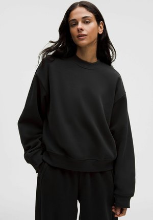 Femme aux longs cheveux foncés portant un sweat-shirt noir oversize et un pantalon noir assorti, debout devant un fond clair uni.