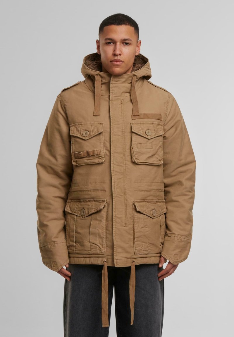 Chaqueta utilitaria color marrón claro con capucha, múltiples bolsillos y bajo con cordón. La tela parece duradera con un acabado texturizado. Diseño simple y práctico.