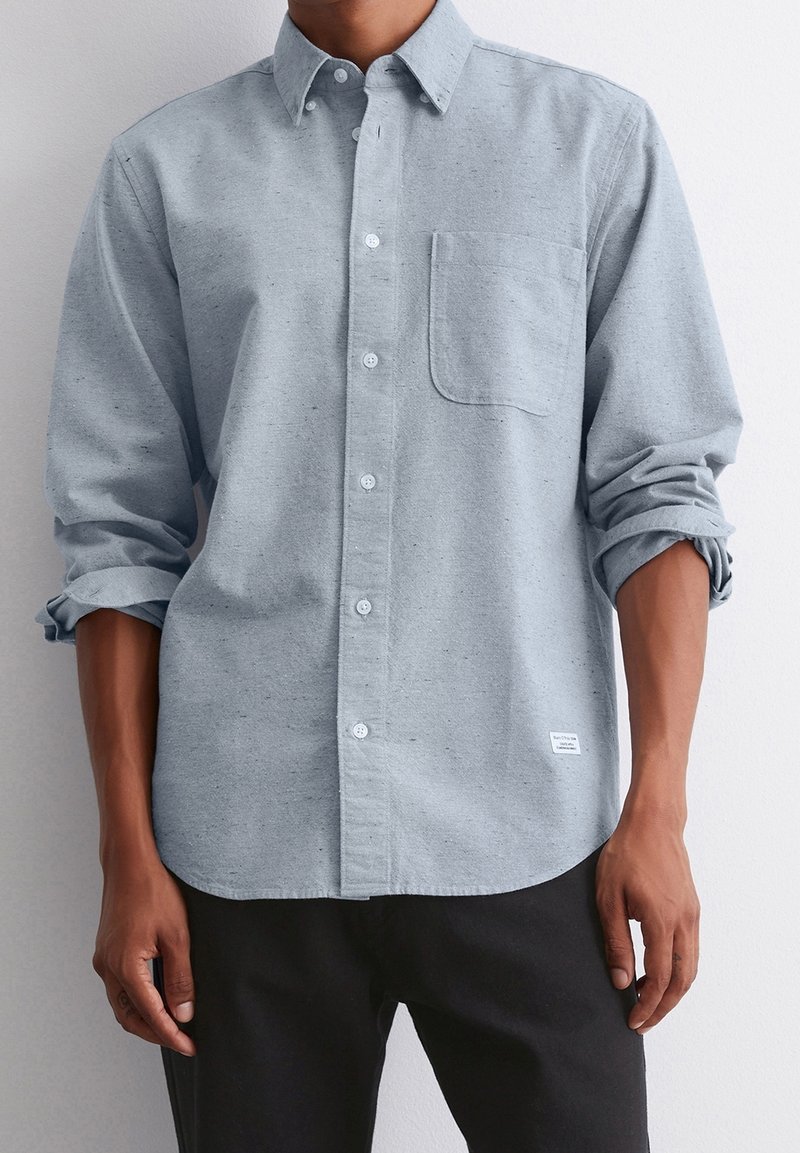 Homme portant une chemise à manches longues gris clair boutonnée avec une poche poitrine et les manches retroussées, associée à un pantalon noir.