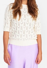 Stickad tröja i krämfärg, med ett mönstrat öppet väv, korta ärmar och ribbat avslut. Matchas med lilafärgad satintights.