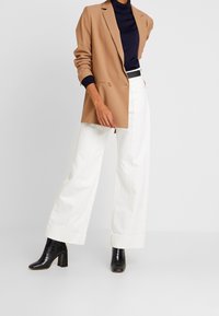 Blazer beige sopra un dolcevita blu navy, abbinato a pantaloni larghi bianchi e stivali neri con tacco. Tessuto liscio con design strutturato.