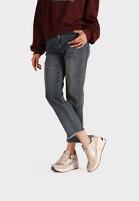 Sneakers beige con una finitura testurizzata, dotati di una suola a zeppa, lacci e accenti metallici, abbinati a jeans in denim a vita alta.
