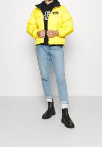 Gul puffjacka med dragkedja och elastiska ärmslut, bärs över en svart hoodie. Ljusblå upprullade jeans och svarta ankelstövlar fulländar outfiten.