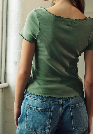 Personne portant une chemise verte à manches courtes avec des bords en dentelle et un jean en denim bleu, se tenant de dos face à l'appareil photo.