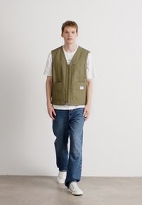 Gilet vert olive avec fermeture éclair à l'avant, deux grandes poches avant, porté sur un T-shirt blanc et un jean bleu, avec des baskets blanches.