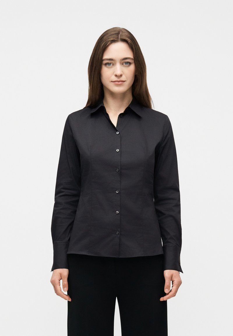 Γυναίκα με ίσια καστανά μαλλιά στέκεται αντικρίζοντας προς τα εμπρός, φορώντας μια εφαρμοστή μαύρη πουκαμίσα με κουμπιά και μαύρο παντελόνι, μπροστά σε ένα απλό φόντο.