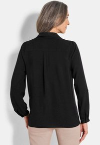 Schwarze Bluse mit langen Ärmeln und Kragen, Rückenschlitzdetail und geknöpften Manschetten. Glattes Material und lockere Passform. Heller Hintergrund.