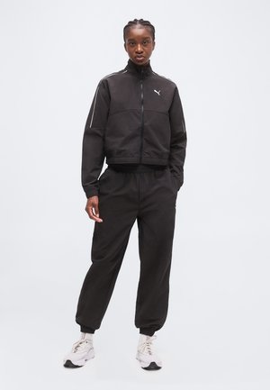 Femme debout portant un survêtement noir Puma avec une veste zippée et un pantalon retroussé, assorti à des baskets blanches sur un fond clair uni.