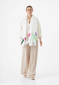 Écharpe blanche avec broderie florale en rose, violet et vert. Portée sur une tenue beige clair, avec un design ample et une texture douce.