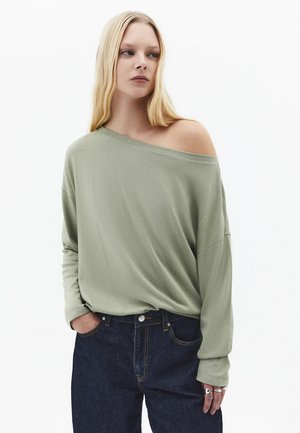 Kvinde med langt blondt hår iført en salviegrøn skulderfri bluse med lange ærmer og mørkeblå jeans, stående med hænderne i lommerne.