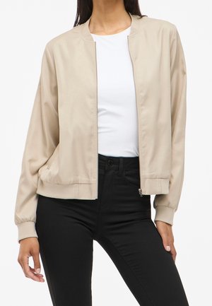 Donna con giacca bomber beige con zip sopra una maglietta bianca e pantaloni neri a vita alta, in piedi in posizione rilassata su sfondo bianco.