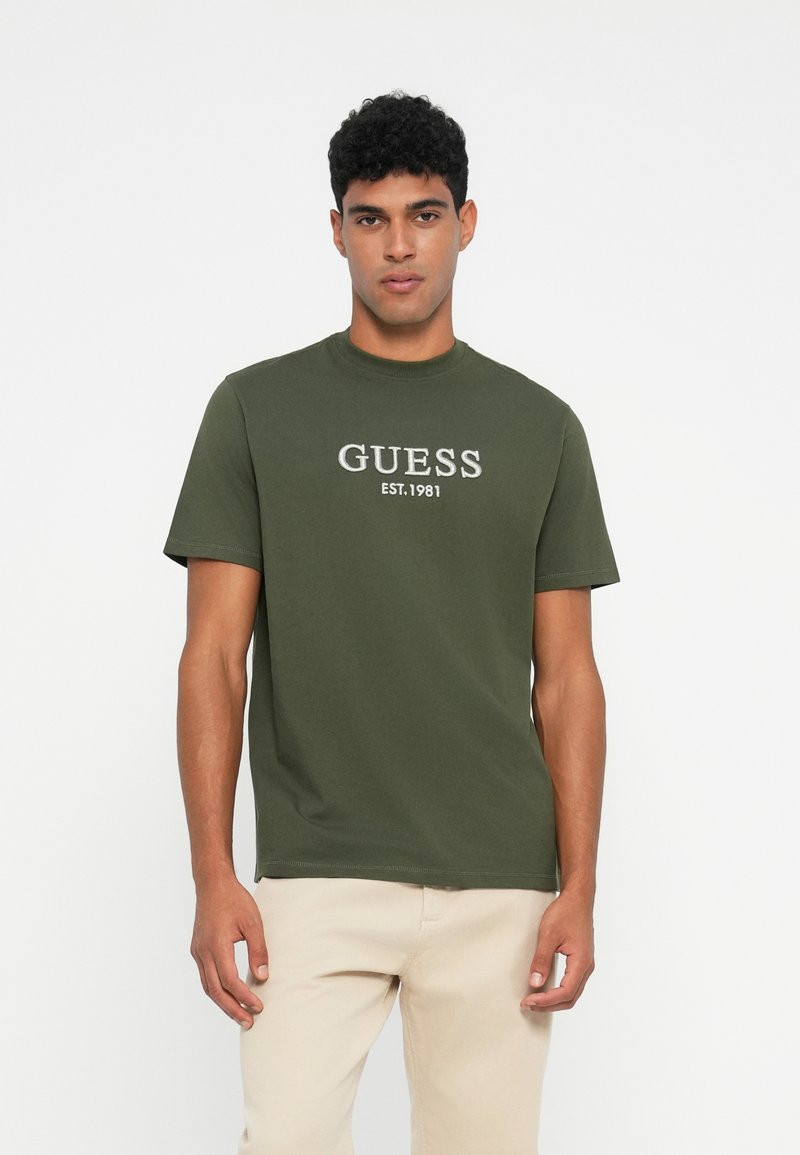 Olijfgroene T-shirt met korte mouwen van katoen, met de tekst "GUESS EST. 1981" in lichtgrijze letters op de borst. Standaard ronde hals.