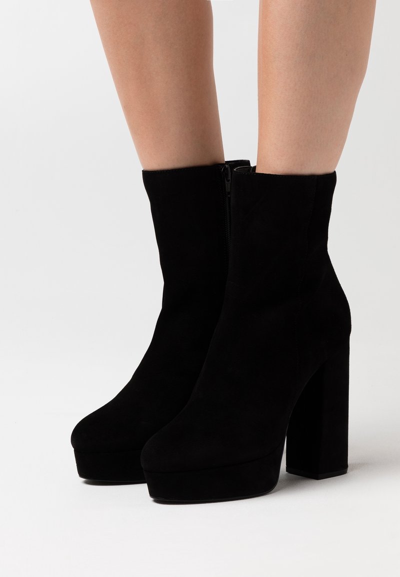Femme portant des bottines à plateforme en daim noir avec talons carrés sur un fond blanc.