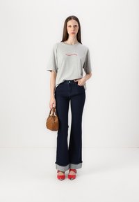 T-shirt en coton gris avec un texte rouge, associé à un jean bleu foncé évasé avec des ourlets en denim, des chaussures à talons rouges et un petit sac à main marron.