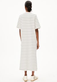 Žena se svázanými kudrnatými vlasy do nízkého drdolu, která má na sobě bílé midi šaty s tenkými černými vodorovnými pruhy a bílé vysoké pletené tenisky, stojící zády směrem k nám.