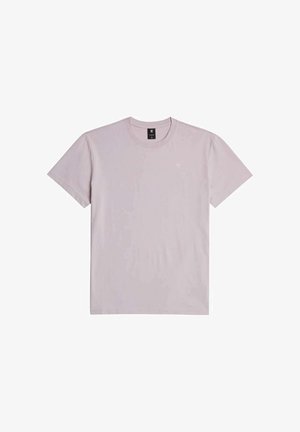 Personne à la peau foncée portant un T-shirt rose clair à manches courtes et un jean gris clair, vue de dos sur un fond uni.
