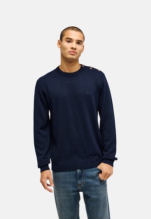 Jeune homme aux cheveux courts portant un pull bleu marine avec des boutons aux épaules et un jean bleu, debout devant un fond clair uni.