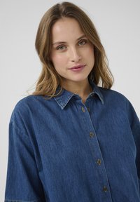 Keskmise sinise denimsärgi ees on nupud, terav krae ja lühikesed varrukad. Omab nähtavat õmblust ja lõdvestatud istuvust.