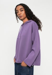 Kobieta z kręconymi włosami, ubrana w luźny fioletowy hoodie i ciemnoniebieskie dżinsy, stojąca z rękami w kieszeniach na jednolitym tle.