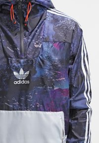 Světle modrá větrovka Adidas s galaktickým vzorem, bílou kapsou na dolní části, třemi bílými pruhy na rukávech a oranžovými šňůrkami na kapuci.