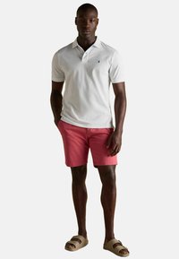 Balsāmīgs balts polo krekls ar īsām piedurknēm, saskaņots ar rozā šortiem. Modelis valkā bēšus sandales. Vienkāršs dizains ar emblēmu uz krūtīm.