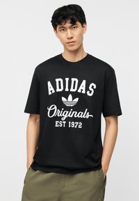 Νεαρός άνδρας που φοράει μαύρο T-shirt Adidas Originals με λευκό λογότυπο και κείμενο, συνδυασμένο με πράσινο-ελαιί παντελόνι, στέκεται μπροστά σε απλό φόντο.