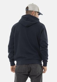 Sweat à capuche bleu marine avec une grande poche au dos et un cordon de serrage réglable. Associé à un pantalon en jean gris et une casquette de couleur claire.