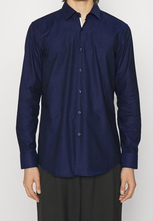 Homme portant une chemise bleu marine à manches longues boutonnée et un pantalon noir, debout devant un fond clair uni.