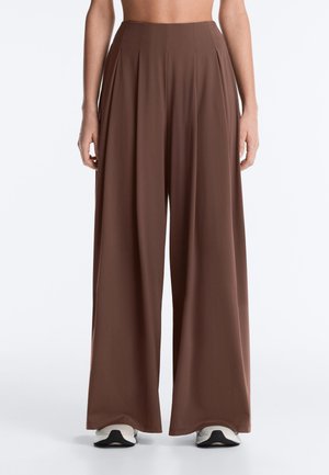 Brązowe spodnie z wysokim stanem i szerokimi nogawkami noszone przez osobę, w połączeniu z białymi i czarnymi trampkami, pokazane od talii do stóp.