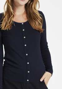 Navy zils kardigans, kas izgatavots no mīksta adījuma materiāla, ar noapaļotu apkakli un sešu gaišu pogu rindu priekšā. Vīļu aproces.
