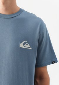 Artimas plano žmogaus, dėvinčio šviesiai mėlyną marškinėlį su ryškiu pilku logotipu krūtinėje, matomas etiketas ant rankovės.