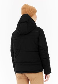 Persoon in een zwarte hoodie, beige gebreide beanie en lichtbruine broek, gezien van achteren tegen een witte achtergrond.